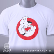 Футболка мисливці за привидами, біла, складена на поверхні, фон світло-сірий, принт з привидом, що тримає червоне сердечко в руках, з етикеткою на зеленій стрічці.