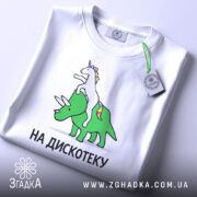 Футболка на дискотеку, біла, зображена на столі з легким нахилом, фон світло-сірий, принт з єдинорогом на трицератопсі, текст "НА ДИСКОТЕКУ" чорним шрифтом, видно етикетку.