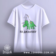 Футболка на дискотеку, біла, підвішена на вішалці, фон світло-сірий, принт з єдинорогом на трицератопсі, текст "НА ДИСКОТЕКУ" чорним шрифтом, видно округлі краї рукавів.
