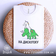 Футболка на дискотеку, біла, складена на плетеному кошику, фон світло-сірий, принт з єдинорогом на трицератопсі, текст "НА ДИСКОТЕКУ" чорним шрифтом, видно етикетку.