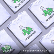 Футболка на дискотеку, біла, зображена на манекені, фон світло-сірий, принт з веселим єдинорогом, що сидить на зеленому трицератопсі, текст "НА ДИСКОТЕКУ" чорним шрифтом.