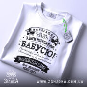 Футболка найкраща бабуся планети, біла, складена на плоскій поверхні, на фоні світлого кольору, принт чорного кольору з текстом на українській мові, з зеленим ярликом.