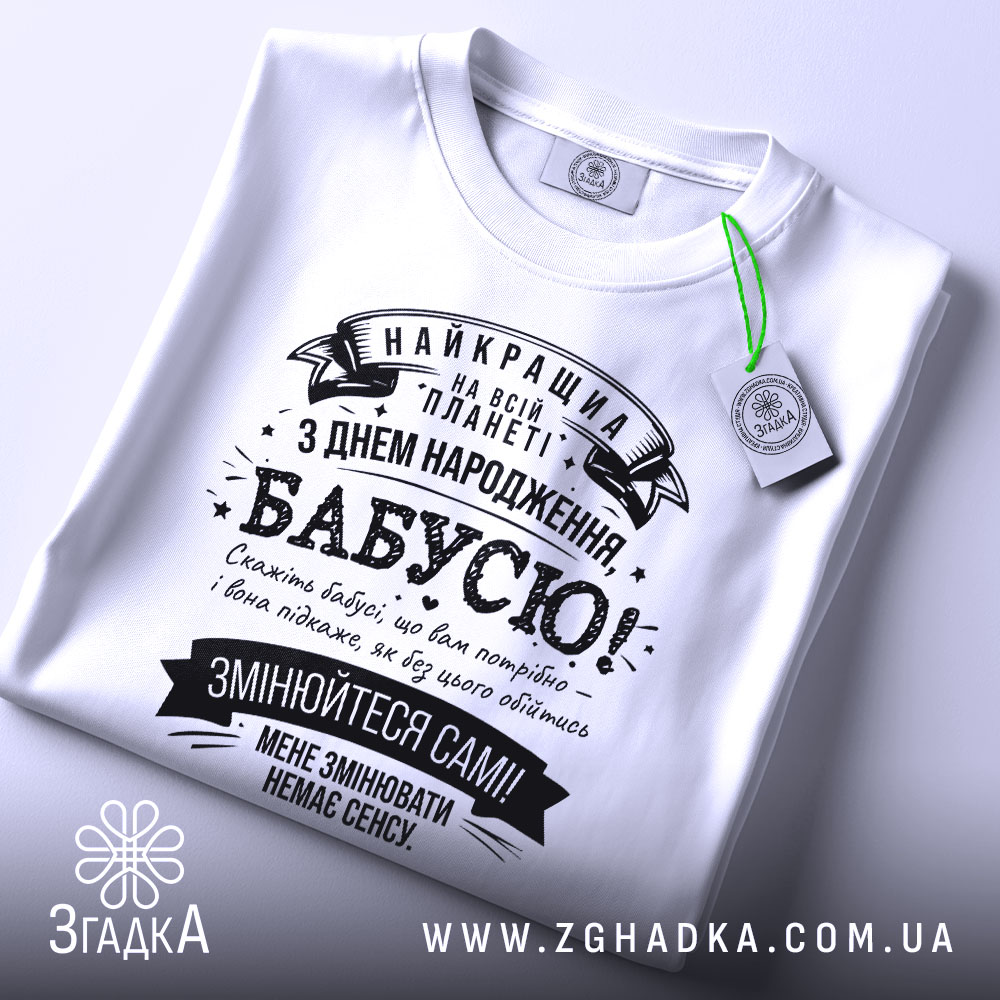 Футболка найкраща бабуся планети, біла, складена на плоскій поверхні, на фоні світлого кольору, принт чорного кольору з текстом на українській мові, з зеленим ярликом.