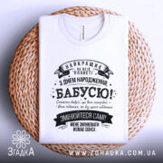 Футболка найкраща бабуся планети, біла, складена на плоскій поверхні з плетеного матеріалу, на фоні світлого кольору, принт чорного кольору з текстом на українській мові.