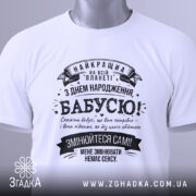 Футболка найкраща бабуся планети, біла, складена на плоскій поверхні, на фоні світлого кольору, принт чорного кольору з текстом на українській мові, з зеленим ярликом.