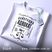 Футболка найкращий адвокат, біла, зображена зверху, складена на поверхні, фон світло-сірий, принт чорного кольору з зображенням ваг та текстом, з етикеткою зеленого кольору.
