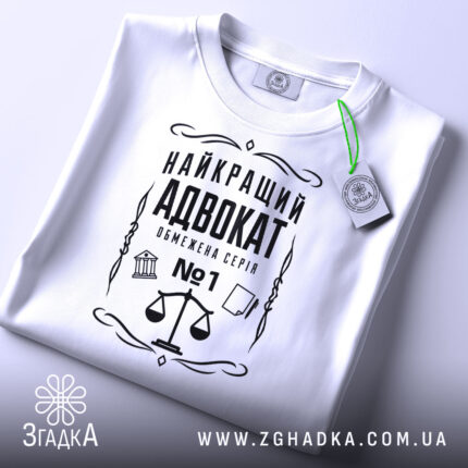 Футболка найкращий адвокат, біла, зображена зверху, складена на поверхні, фон світло-сірий, принт чорного кольору з зображенням ваг та текстом, з етикеткою зеленого кольору.