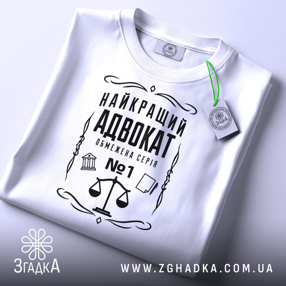 Футболка найкращий адвокат, біла, зображена зверху, складена на поверхні, фон світло-сірий, принт чорного кольору з зображенням ваг та текстом, з етикеткою зеленого кольору.