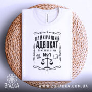 Футболка найкращий адвокат, біла, зображена зверху, складена на плетеній кошику, фон світло-сірий, принт чорного кольору з зображенням ваг та текстом.