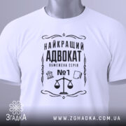 Футболка найкращий адвокат, біла, зображена зверху, складена на поверхні, фон світло-сірий, принт чорного кольору з зображенням ваг та текстом, з етикеткою зеленого кольору.