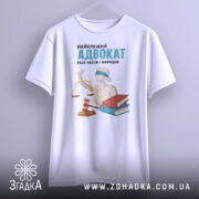 Футболка найкращий адвокат, біла, зображена з фронтального ракурсу, фон світло-сірий, принт містить зображення статуї правосуддя, червоного молотка та двох книг, напис великими літерами: 'НАЙКРАЩИЙ АДВОКАТ УСІХ ЧАСІВ І НАРОДІВ'.