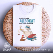 Футболка найкращий адвокат, біла, складена на плоскій поверхні, фон плетений, принт містить зображення статуї правосуддя, червоного молотка та двох книг, напис: 'НАЙКРАЩИЙ АДВОКАТ УСІХ ЧАСІВ І НАРОДІВ'.