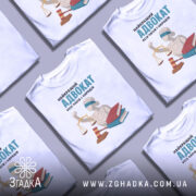 Футболка найкращий адвокат, біла, зображена з фронтального ракурсу, на фоні світло-сірого кольору, принт містить зображення статуї правосуддя з вагою, червоного молотка та двох книг, напис великими літерами: 'НАЙКРАЩИЙ АДВОКАТ УСІХ ЧАСІВ І НАРОДІВ'.