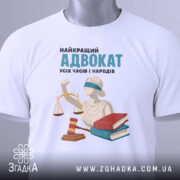 Футболка найкращий адвокат, біла, складена на плоскій поверхні, фон світло-сірий, принт зображує статую правосуддя, червоний молоток та дві книги, напис: 'НАЙКРАЩИЙ АДВОКАТ УСІХ ЧАСІВ І НАРОДІВ', на футболці є зелений етикет.