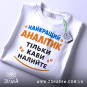 Футболка найкращий аналітик, білого кольору, розкладена на поверхні, фон світло-сірий, принт з текстом синього та оранжевого кольорів, з етикеткою на зеленій стрічці.