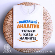 Футболка найкращий аналітик, білого кольору, розкладена на плетеній кошику, фон білий, принт з текстом синього та оранжевого кольорів, з кавовими зернами.