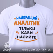 Футболка найкращий аналітик, білого кольору, розкладена на поверхні, фон світло-сірий, принт з текстом синього та оранжевого кольорів, з етикеткою на зеленій стрічці.