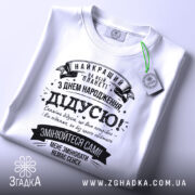 Футболка найкращий дідусь планети, біла, лежить на поверхні, фон світлий, принт чорного кольору з текстом на українській мові, прикрашений стрічками та зірочками, на комірці видна етикетка.