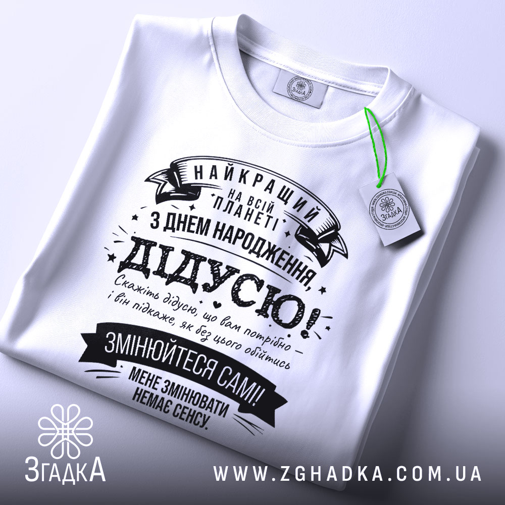 Футболка найкращий дідусь планети, біла, лежить на поверхні, фон світлий, принт чорного кольору з текстом на українській мові, прикрашений стрічками та зірочками, на комірці видна етикетка.