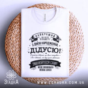 Футболка найкращий дідусь планети, біла, лежить на плетеній кошику, фон натуральний, принт чорного кольору з текстом на українській мові, оформлений у стилі ретро з декоративними елементами.