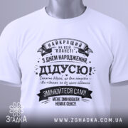 Футболка найкращий дідусь планети, біла, лежить на поверхні, фон світлий, принт чорного кольору з текстом на українській мові, прикрашений стрічками та зірочками, на комірці видна етикетка.
