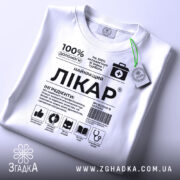 Футболка найкращий лікар, біла, складена на столі, фон світло-сірий, принт чорного кольору з медичними символами, на комірці зелений ярлик.