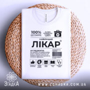 Футболка найкращий лікар, біла, складена на плетеній тарілці, фон світло-сірий, принт чорного кольору з медичними символами та текстом.