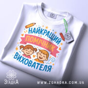 Футболка найкращий помічник вихователя, білого кольору, складена на столі, фон світло-сірий, принт з малюнками дітей, олівець та зірочки, на футболці є етикетка з зеленим шнурком.