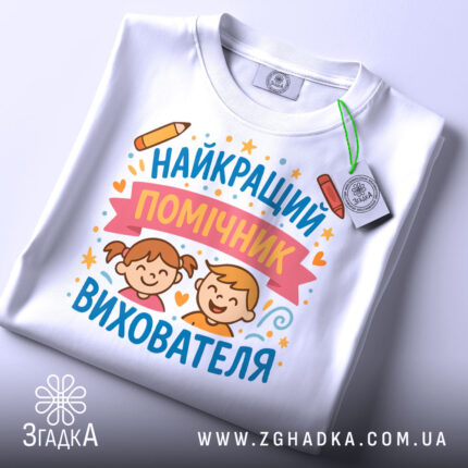 Футболка найкращий помічник вихователя, білого кольору, складена на столі, фон світло-сірий, принт з малюнками дітей, олівець та зірочки, на футболці є етикетка з зеленим шнурком.