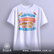 Футболка найкращий помічник вихователя, білого кольору, підвішена на вішалці, фон світло-сірий, принт з малюнками дітей, олівці та зірочки навколо тексту.