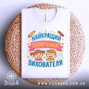 Футболка найкращий помічник вихователя, білого кольору, складена на плетеному кошику, фон світло-сірий, принт з малюнками дітей, олівець та зірочки, видно етикетку на горловині.