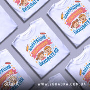 Футболка найкращий помічник вихователя, білого кольору, зображена на манекені, фон світло-сірий, принт містить малюнки двох дітей з усмішками, олівці та зірочки навколо тексту.