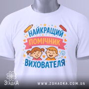 Футболка найкращий помічник вихователя, білого кольору, складена на столі, фон світло-сірий, принт з малюнками дітей, олівець та зірочки, на футболці є етикетка з зеленим шнурком.