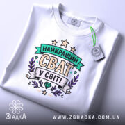 Футболка найкращий сват, біла, складена на плоскій поверхні, фон світло-сірий, принт з написом 'НАЙКРАЩИЙ СВАТ У СВІТІ' в кольорах зеленого, фіолетового та жовтого, з етикеткою на зеленому шнурку.