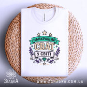 Футболка найкращий сват, біла, складена на плоскій поверхні, фон білий, принт з написом 'НАЙКРАЩИЙ СВАТ У СВІТІ' в кольорах зеленого, фіолетового та жовтого, на плетеній основі.