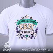 Футболка найкращий сват, біла, складена на плоскій поверхні, фон світло-сірий, принт з написом 'НАЙКРАЩИЙ СВАТ У СВІТІ' в кольорах зеленого, фіолетового та жовтого, з етикеткою на зеленому шнурку.