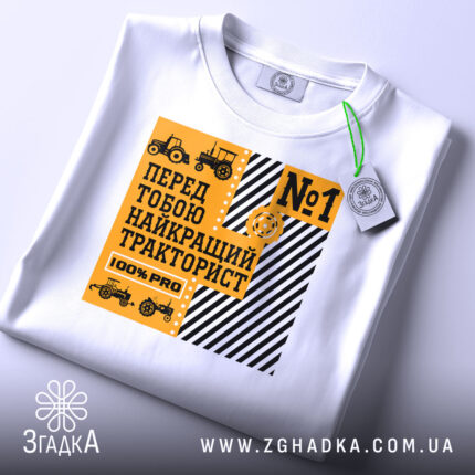 Футболка найкращий тракторист. Біла футболка, складена на поверхні, з видимим принтом. На фоні світло-сірий колір. Принт жовтого кольору з чорними елементами, текст 'Найкращий тракторист' та зображення тракторів. На футболці є етикетка з зеленим шнурком.