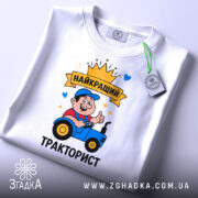 Футболка найкращий тракторист. Біла футболка, складена на столі, принт з трактористом, на футболці є етикетка з зеленим шнурком, фон світло-сірий.
