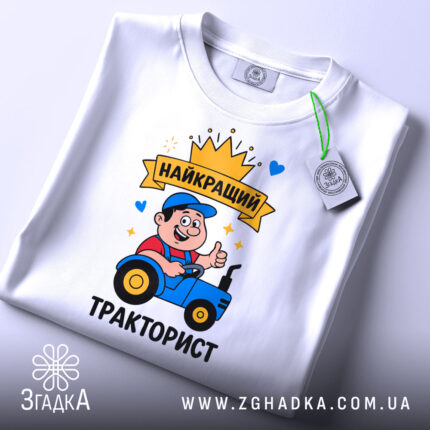 Футболка найкращий тракторист. Біла футболка, складена на столі, принт з трактористом, на футболці є етикетка з зеленим шнурком, фон світло-сірий.
