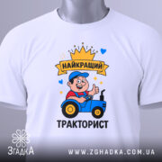 Футболка найкращий тракторист. Біла футболка, складена на столі, принт з трактористом, на футболці є етикетка з зеленим шнурком, фон світло-сірий.