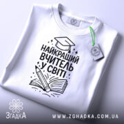 Футболка найкращий вчитель, білого кольору, зображена зверху, на столі, фон світло-сірий, принт чорного кольору з малюнком книги та олівця, на футболці є етикетка.
