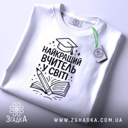 Футболка найкращий вчитель, білого кольору, зображена зверху, на столі, фон світло-сірий, принт чорного кольору з малюнком книги та олівця, на футболці є етикетка.