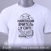Футболка найкращий вчитель, білого кольору, зображена зверху, на столі, фон світло-сірий, принт чорного кольору з малюнком книги та олівця, на футболці є етикетка.
