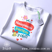 Футболка найкращий вчитель, біла, складена на поверхні, фон світло-сірий, принт яскравий з текстом та елементами декору, прикріплена етикетка.