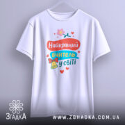 Футболка найкращий вчитель, біла, підвішена на вішалці, фон світло-сірий, принт кольоровий з текстом та ілюстраціями, виглядає стильно.