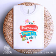 Футболка найкращий вчитель, біла, складена на плетеній основі, фон світло-сірий, принт яскравий з текстом та квітами, виглядає акуратно.