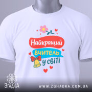 Футболка найкращий вчитель, біла, складена на поверхні, фон світло-сірий, принт яскравий з текстом та елементами декору, прикріплена етикетка.