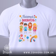 Футболка найкращий вихователь, білого кольору, складена на поверхні, фон світлий, принт зображує чотирьох дітей, кольорові елементи, такі як цукерки та квіти, а також етикетка з зеленим шнурком.