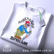 Футболка Найкращий зварювальник, білого кольору, зображена на столі з верхнього ракурсу, фон світло-сірий, принт з малюнком зварювальника у синій куртці та червоній касці, що працює з електродом, текст на футболці чорний, на футболці є етикетка.