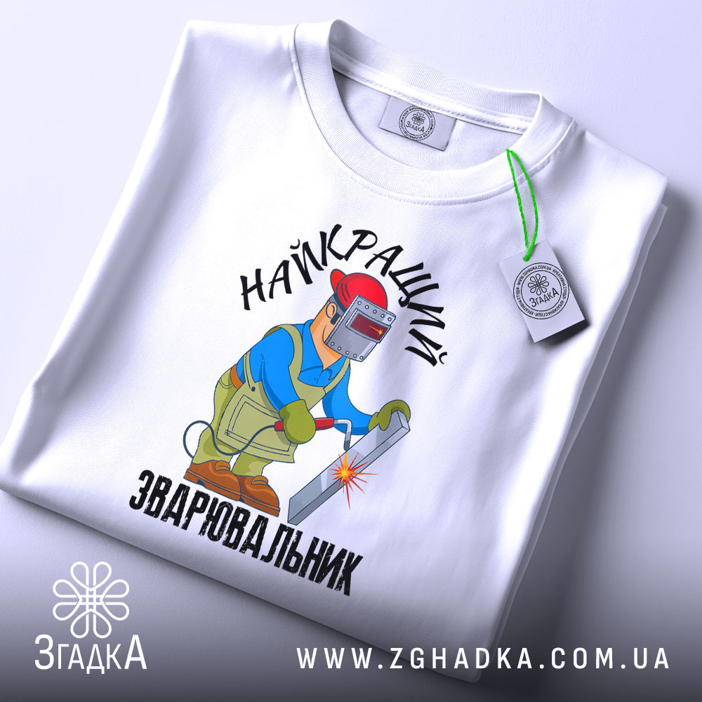 Футболка Найкращий зварювальник, білого кольору, зображена на столі з верхнього ракурсу, фон світло-сірий, принт з малюнком зварювальника у синій куртці та червоній касці, що працює з електродом, текст на футболці чорний, на футболці є етикетка.